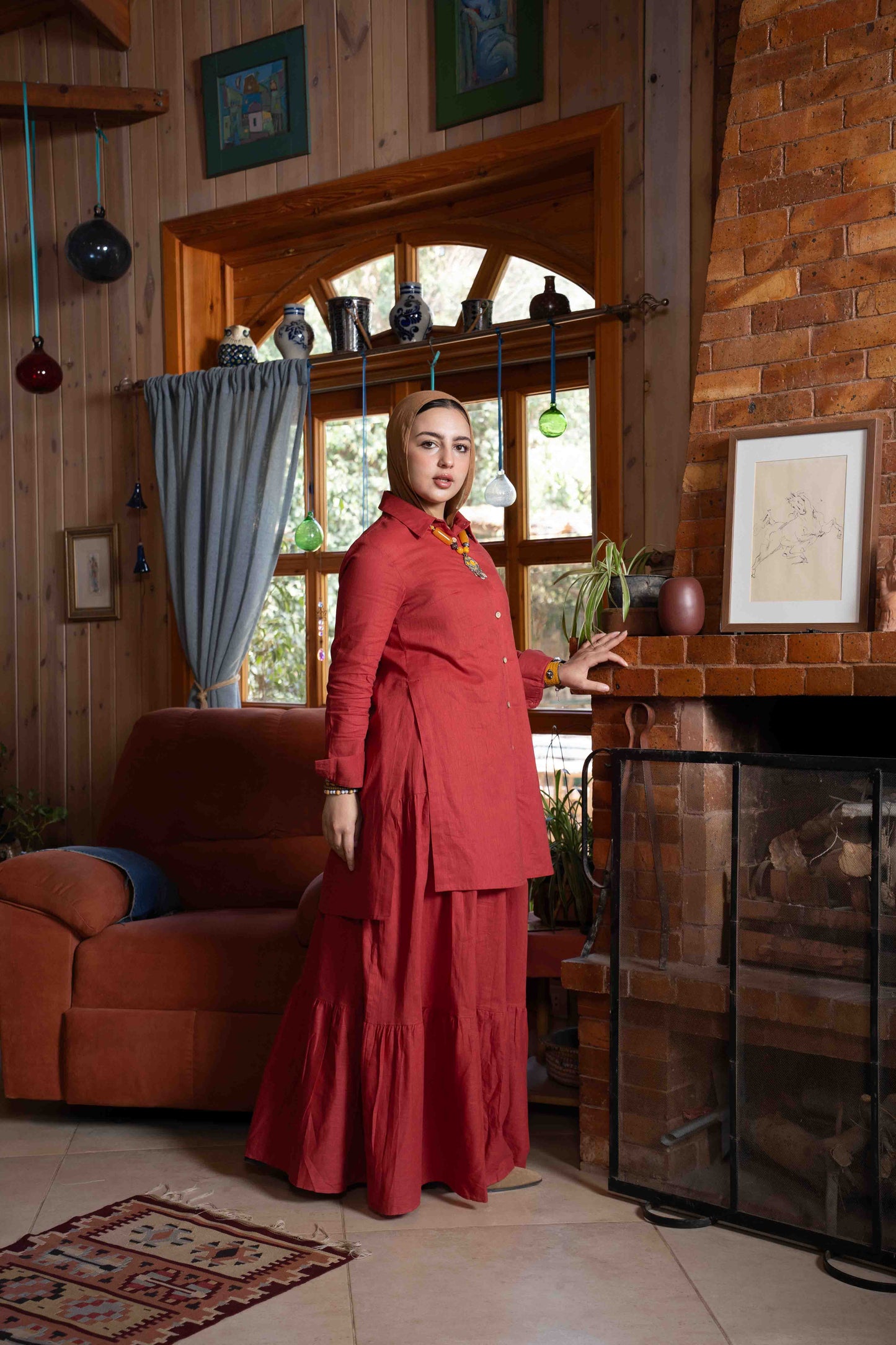 Woman in a red outfit standing in a cozy room with wooden walls and a fireplace.
موديل ترتدي طقم تنورة زهرة باللون العنابي الأنيق من براند Kaleido