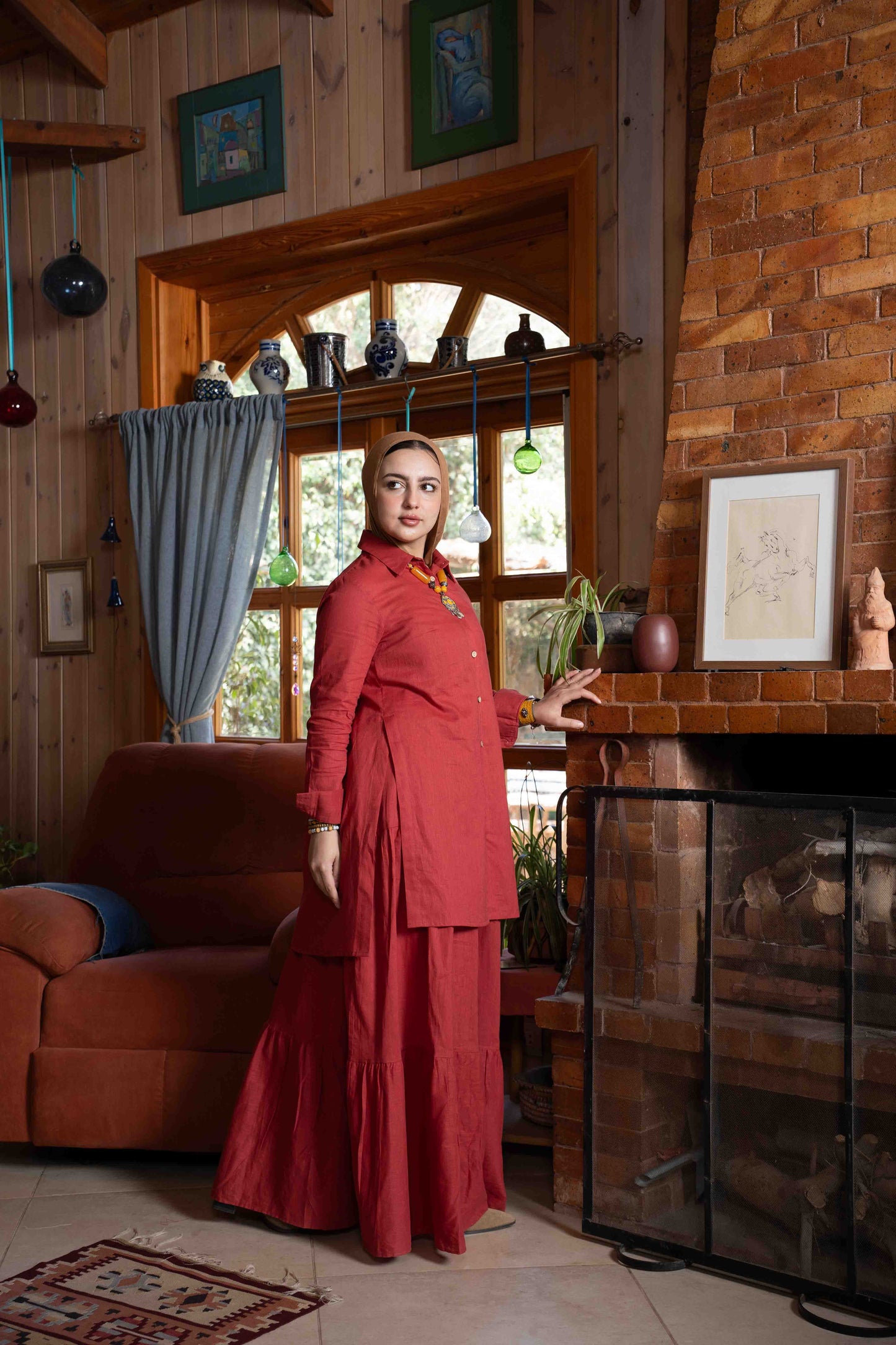 Woman in a red outfit standing in a cozy room with wooden walls and a fireplace.
موديل ترتدي طقم تنورة زهرة باللون العنابي الأنيق من براند Kaleido