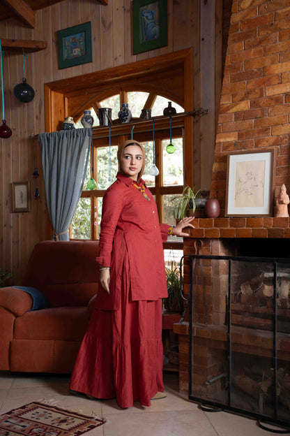 Woman in a red outfit standing in a cozy room with wooden walls and a fireplace.
موديل ترتدي طقم تنورة زهرة باللون العنابي الأنيق من براند Kaleido