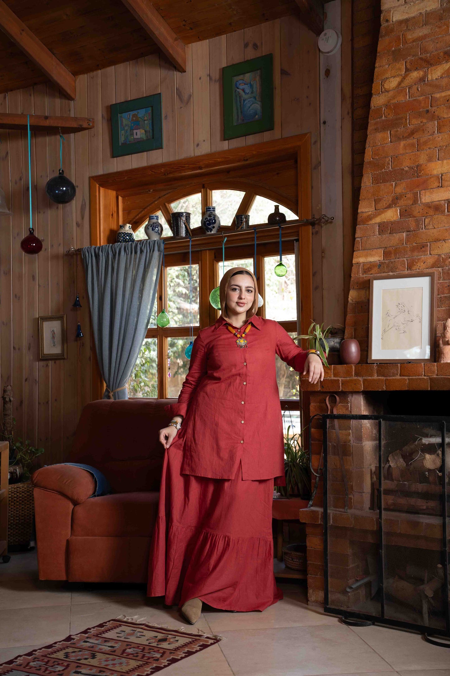 Woman in a red outfit standing in a cozy room with wooden walls and a fireplace.
موديل ترتدي طقم تنورة زهرة باللون العنابي الأنيق من براند Kaleido