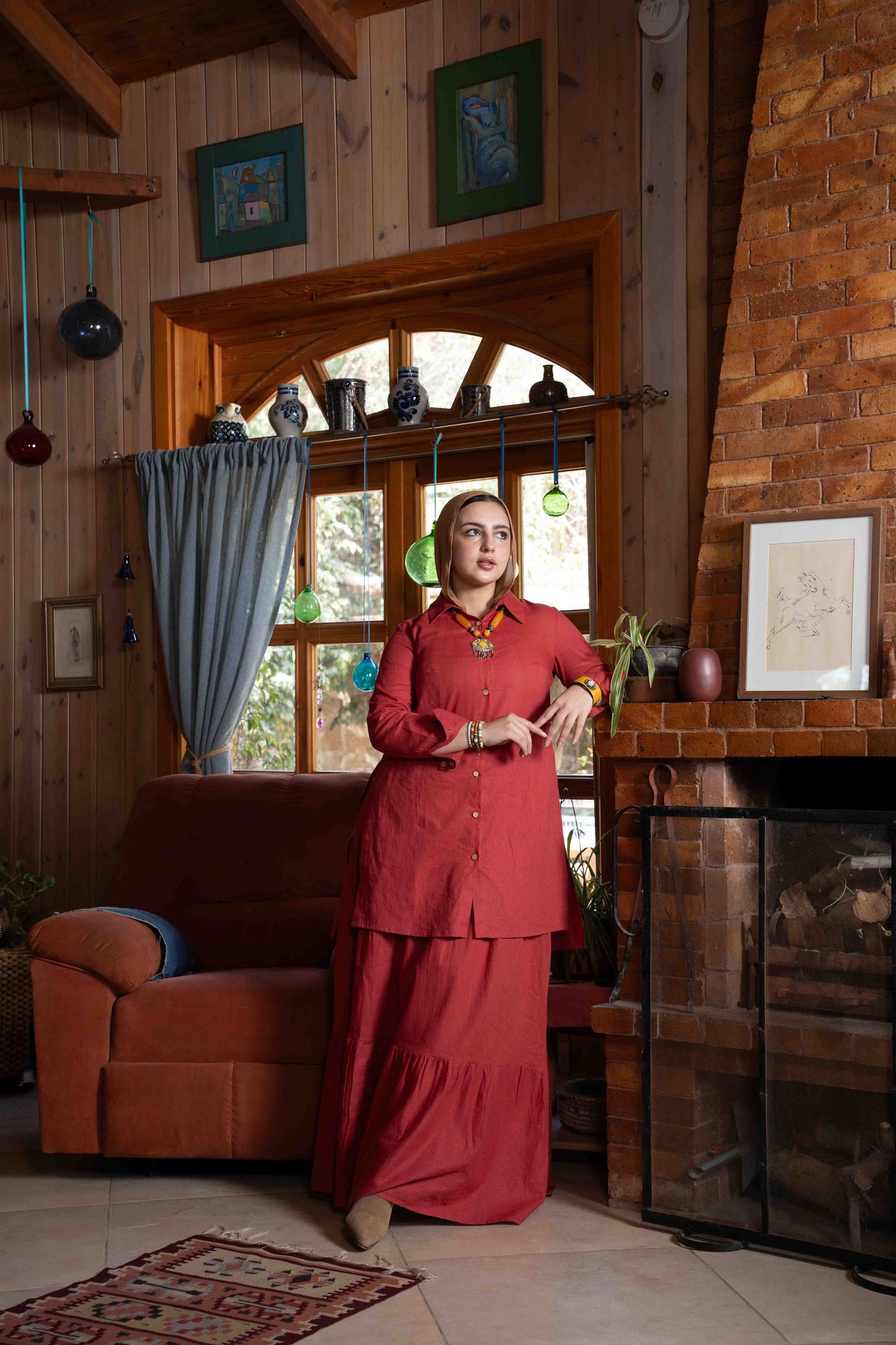 Woman in a red outfit standing in a cozy living room with wooden walls and a fireplace. بنت محجبة ترتدي شميز واسع محتشم و جيبة واسعة بلون احمر