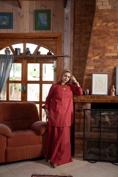 Woman in a red outfit standing in a cozy room with wooden walls and a fireplace.
موديل ترتدي طقم تنورة زهرة باللون العنابي الأنيق من براند Kaleido