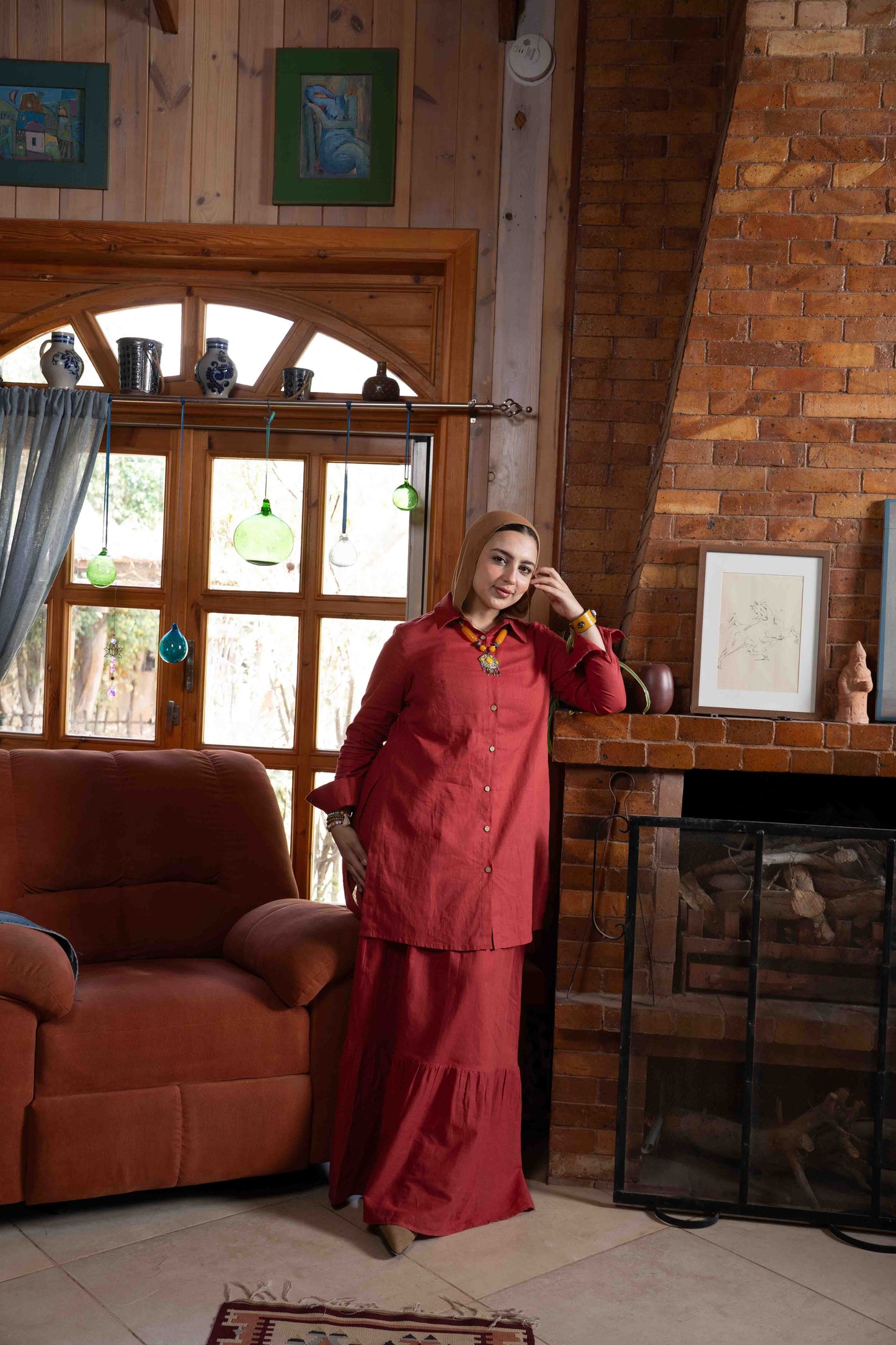 Woman in a red outfit standing in a cozy living room with wooden walls and a fireplace. بنت محجبة ترتدي شميز واسع محتشم و جيبة واسعة بلون احمر
