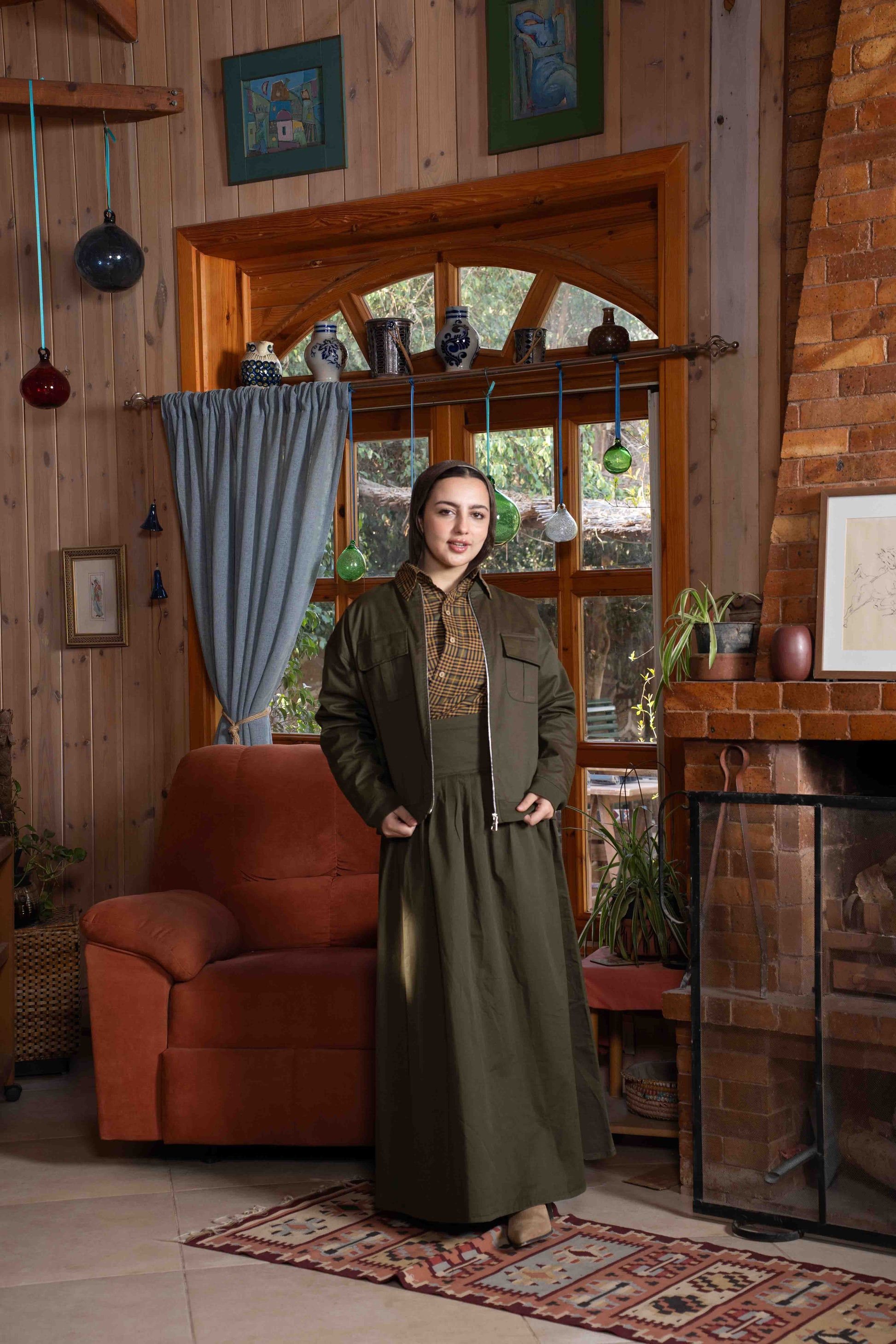 Woman in a long green dress standing in a cozy living room with wooden walls and a fireplace.