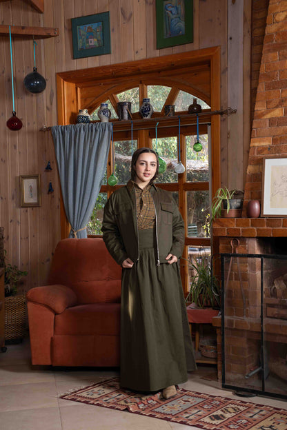 Woman in a long green dress standing in a cozy living room with wooden walls and a fireplace.